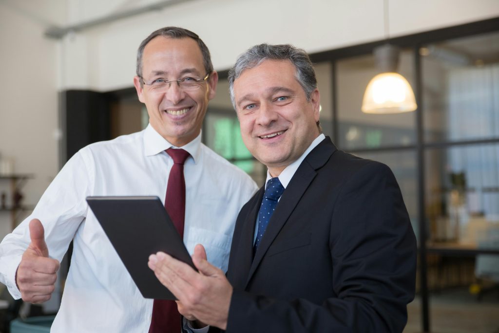 A man in a black suit wearing a blue tie with white polka dots, holding a tablet standing beside a man in a white shirt holding a thumb up