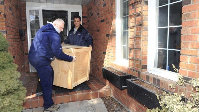 Two people moving a large wooden crate up steps to a house.